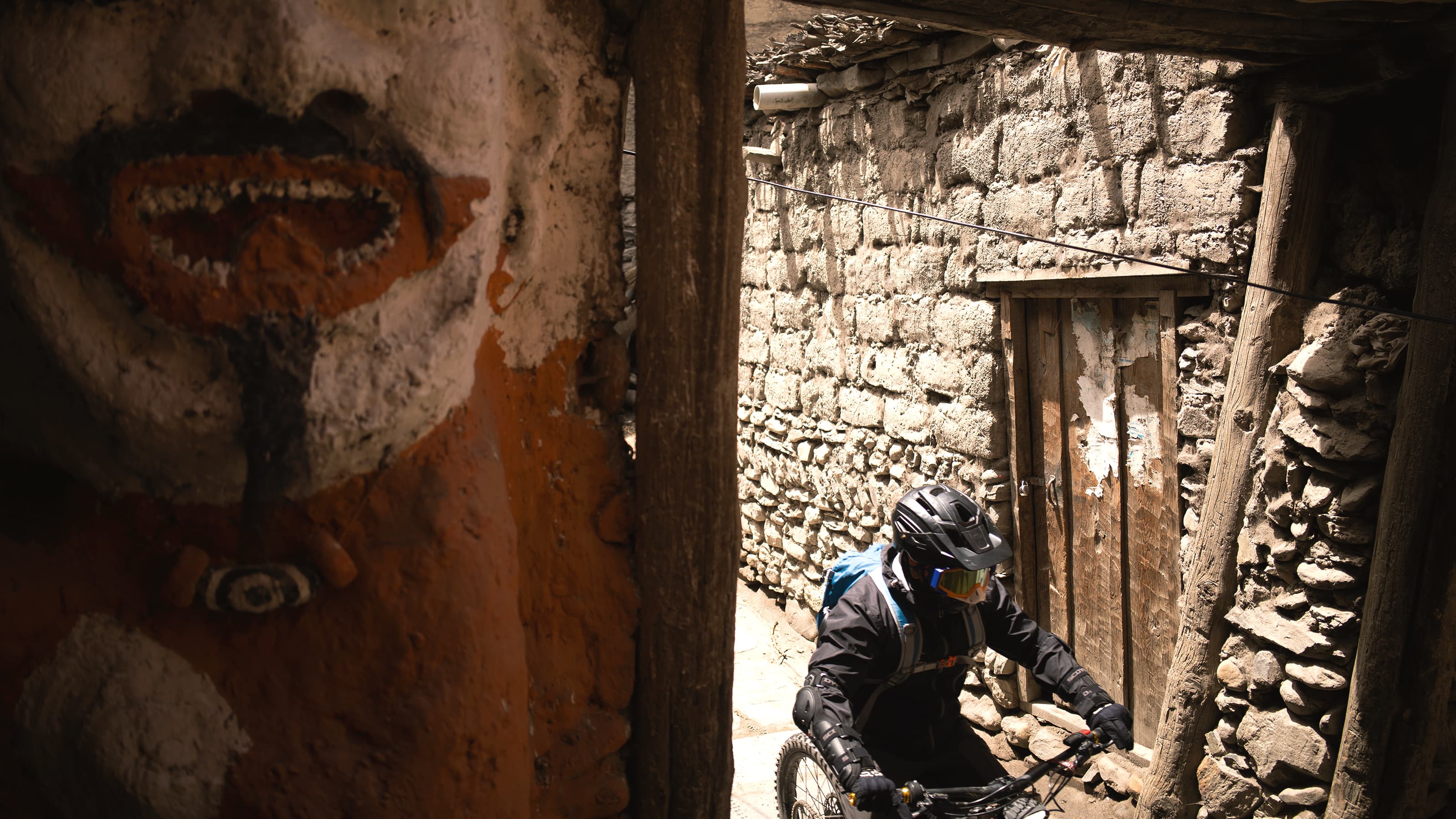 E-bike rider passing a painted guardian mask in a narrow stone alley