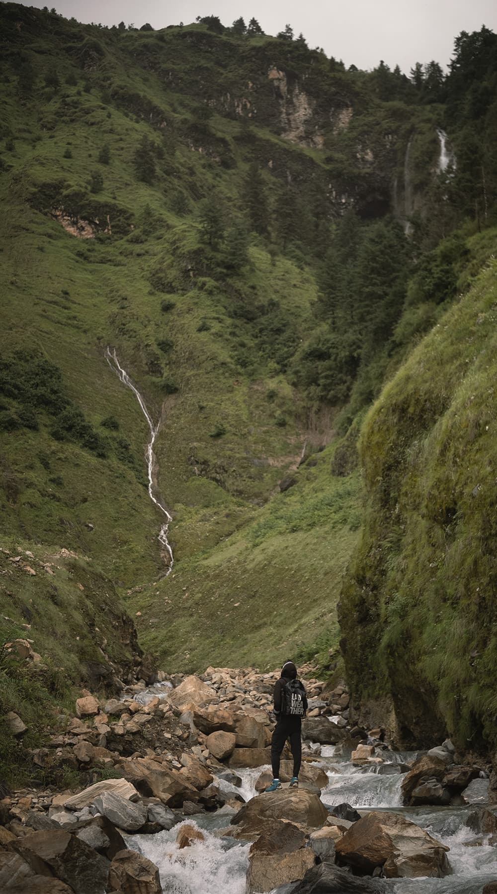 Hiker standing on boulders in a stream below grassy cliffs and waterfalls