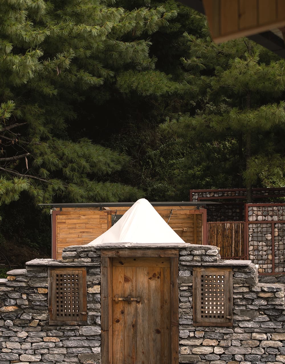 White tent peak rising behind a stone wall and timber door among pines